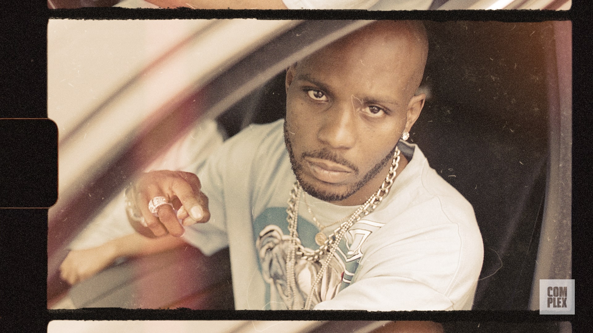 A man with a serious expression, wearing a chain necklace and a graphic t-shirt, sits in a car, looking directly at the camera.