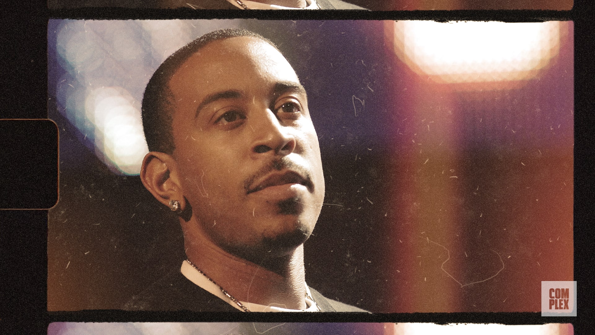 Ludacris, wearing earrings and a chain, looks thoughtful against a colorful, blurred background.