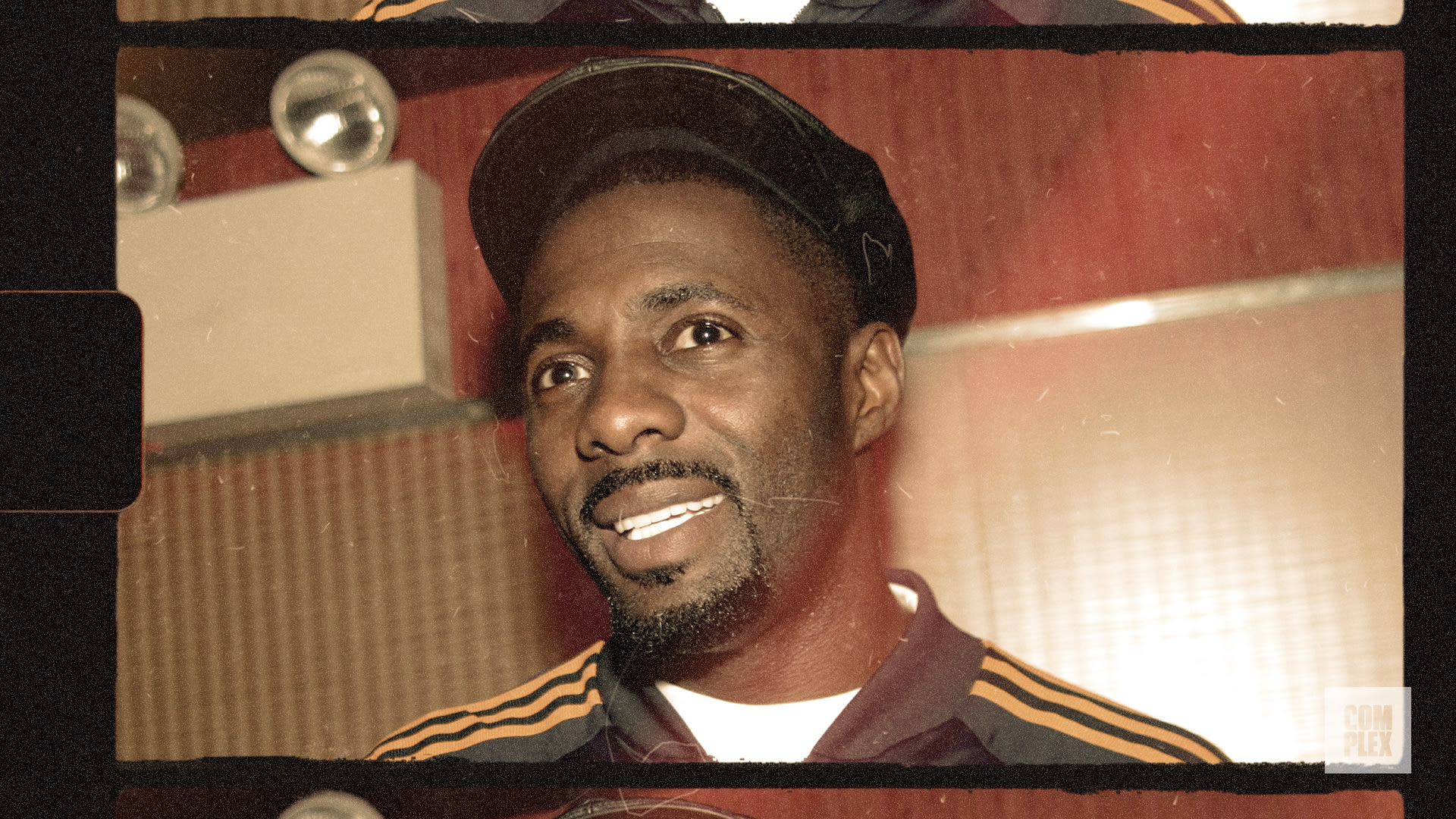 A man wearing a cap and a sports jacket with stripes, smiling in a warmly lit room.