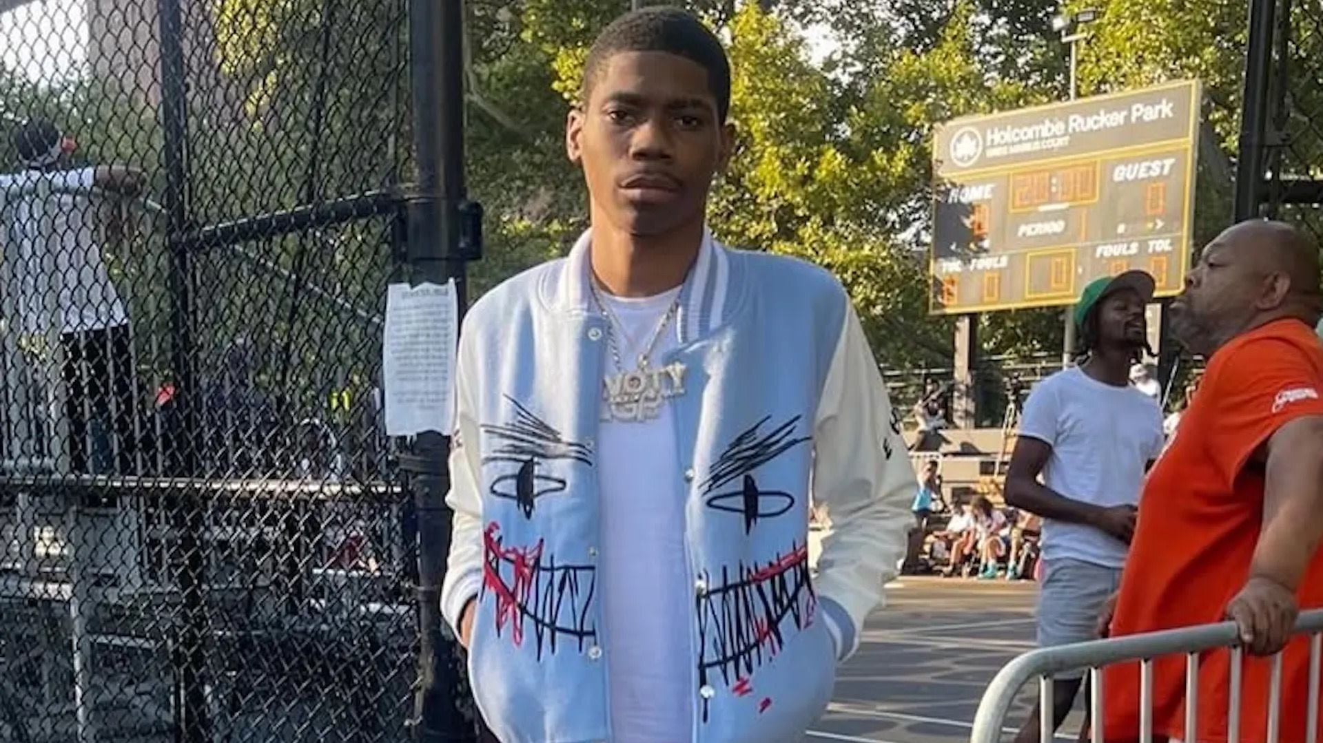 A man in a blue and white jacket stands near a basketball court at Holcombe Rucker Park, with people and a scoreboard in the background.