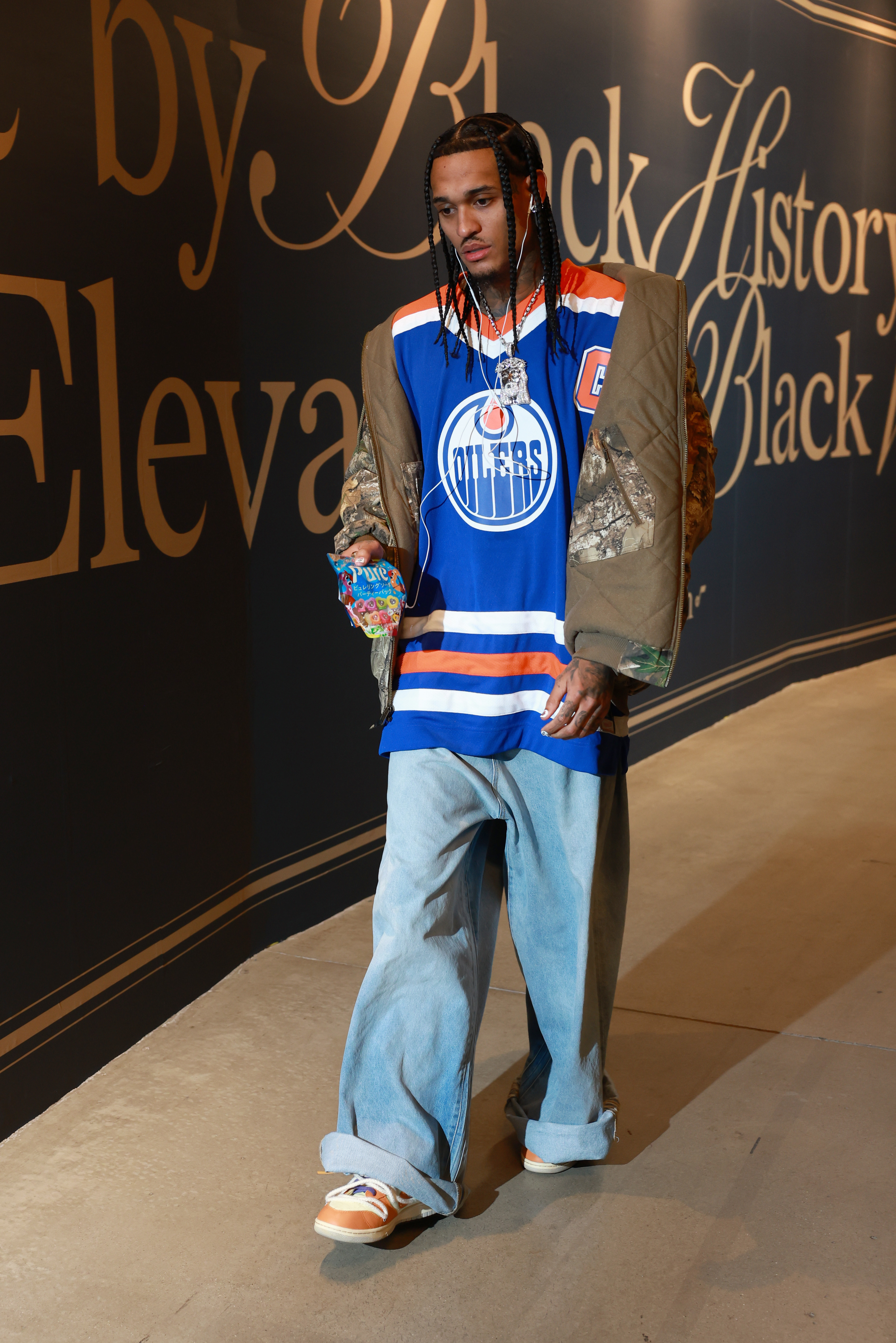 A person in a blue Oilers jersey, camo jacket, and baggy jeans walks indoors. They have braided hair and hold colorful candy.