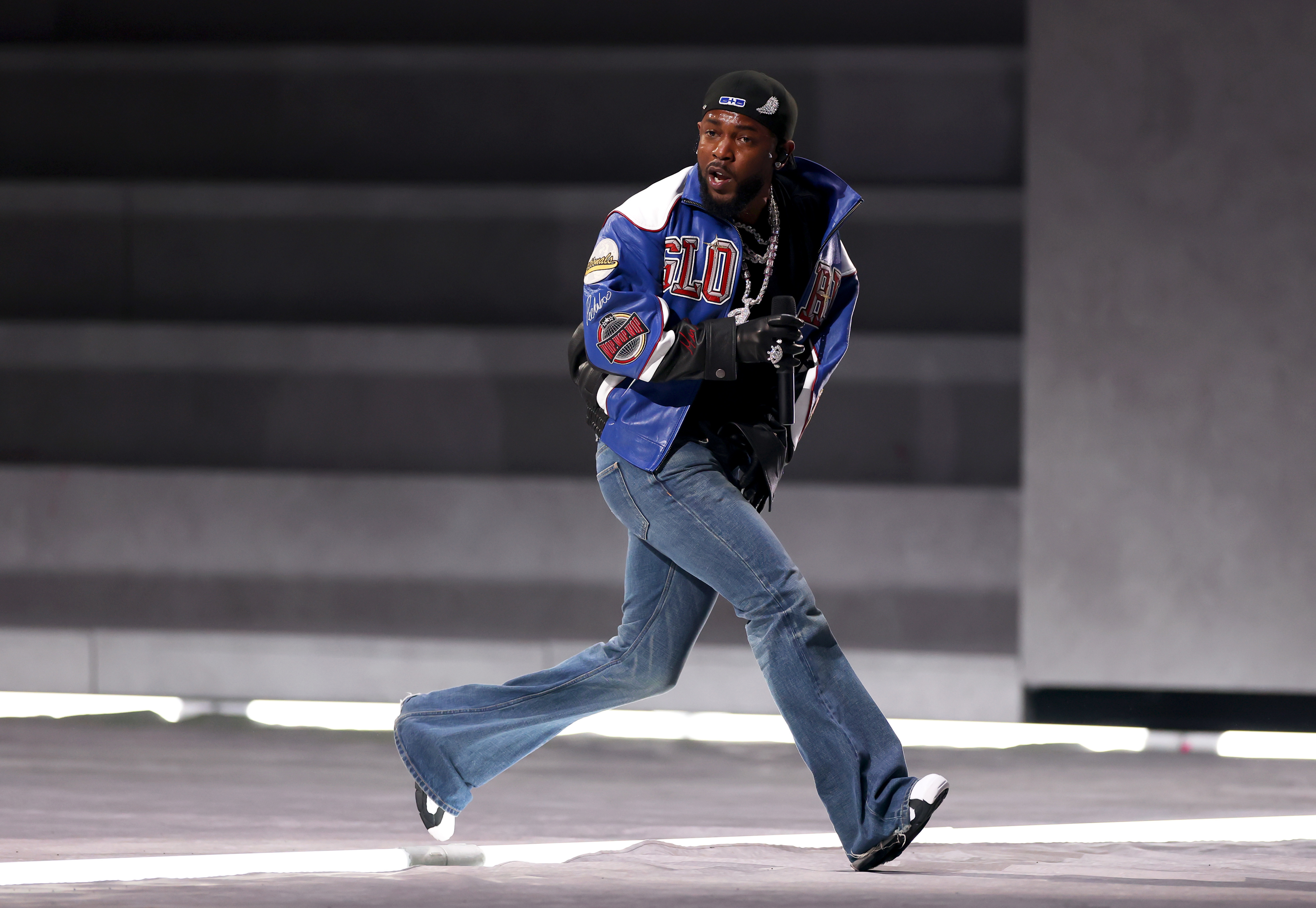 Kendrick Lamar is energetically running on a stage, wearing a colorful jacket, jeans, and a cap during the Super Bowl. He will be up for a best rap grammy. 