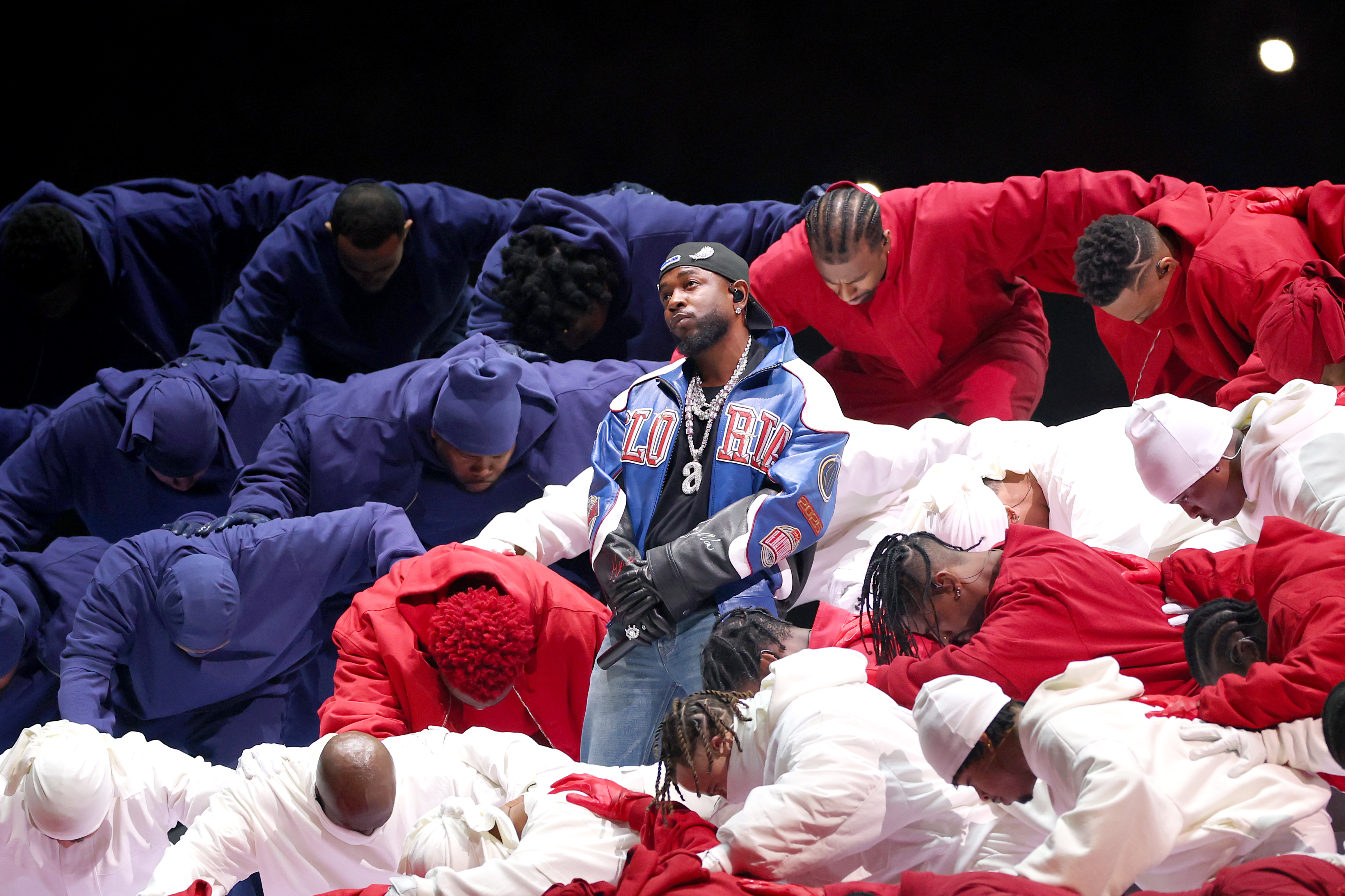 Kendrick Lamar a blue and white jacket playing the Super Bowl. He is standing amidst dancers in red, white, and blue outfits, who are bent forward. He might win a Grammy in 2026. 