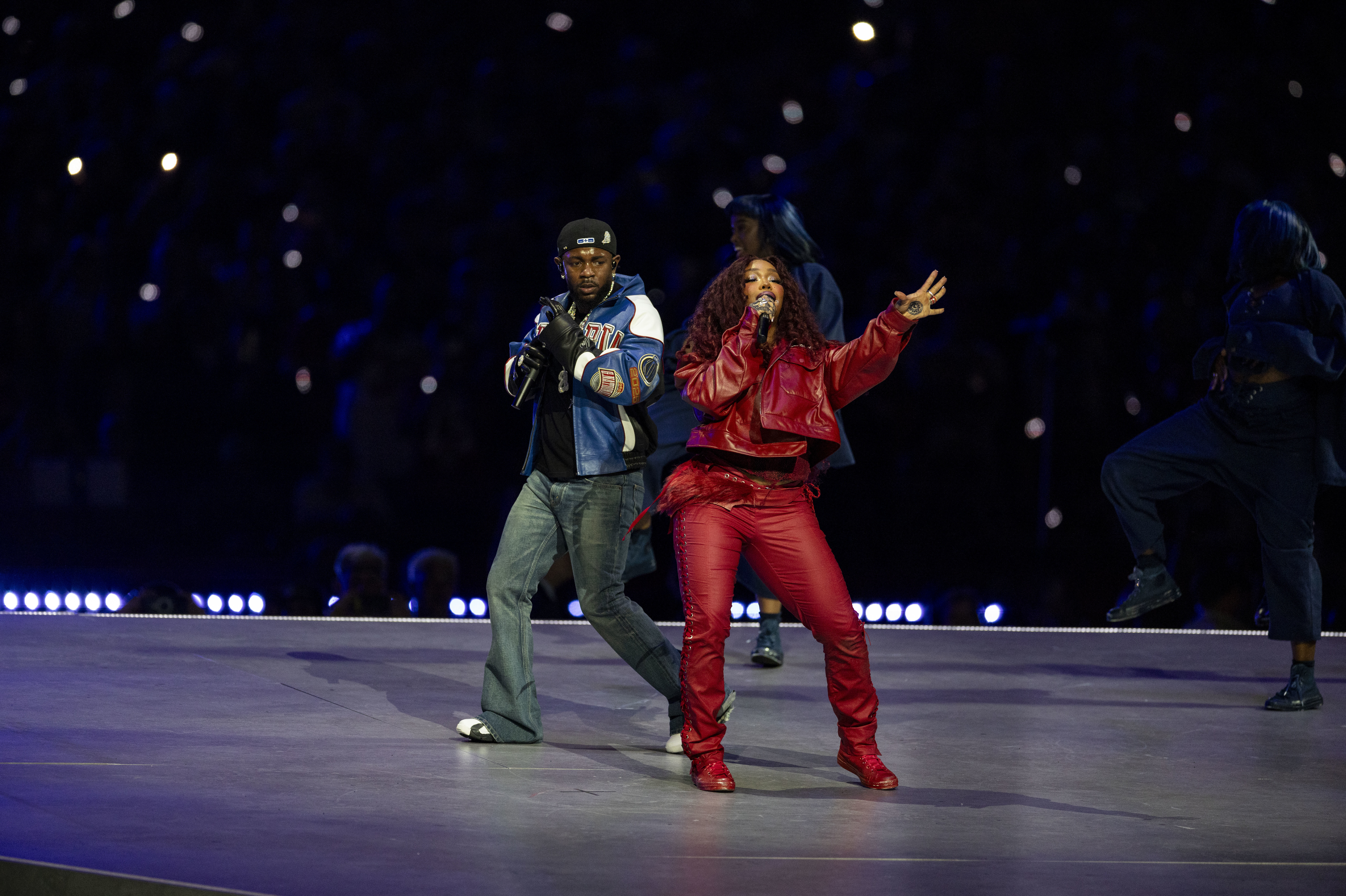 SZA, n a red outfit sings energetically on stage with Kendrick Lamar in a blue jacket and jeans. The background is dimly lit. Their song 