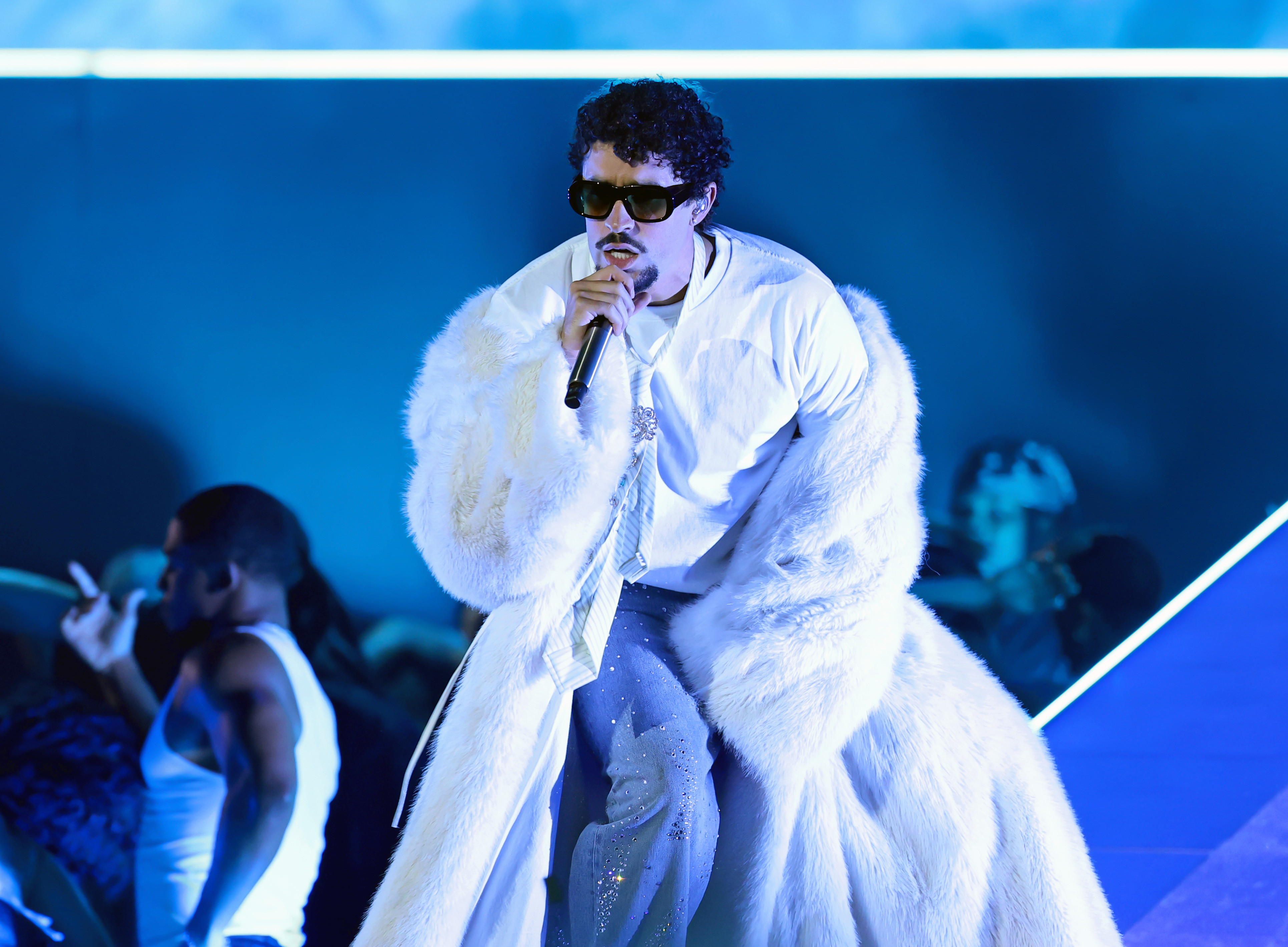 Bad Bunny in a white fur coat and sunglasses sings on stage, with a blue-lit background. He will be a big winner at the 2026 Grammys.