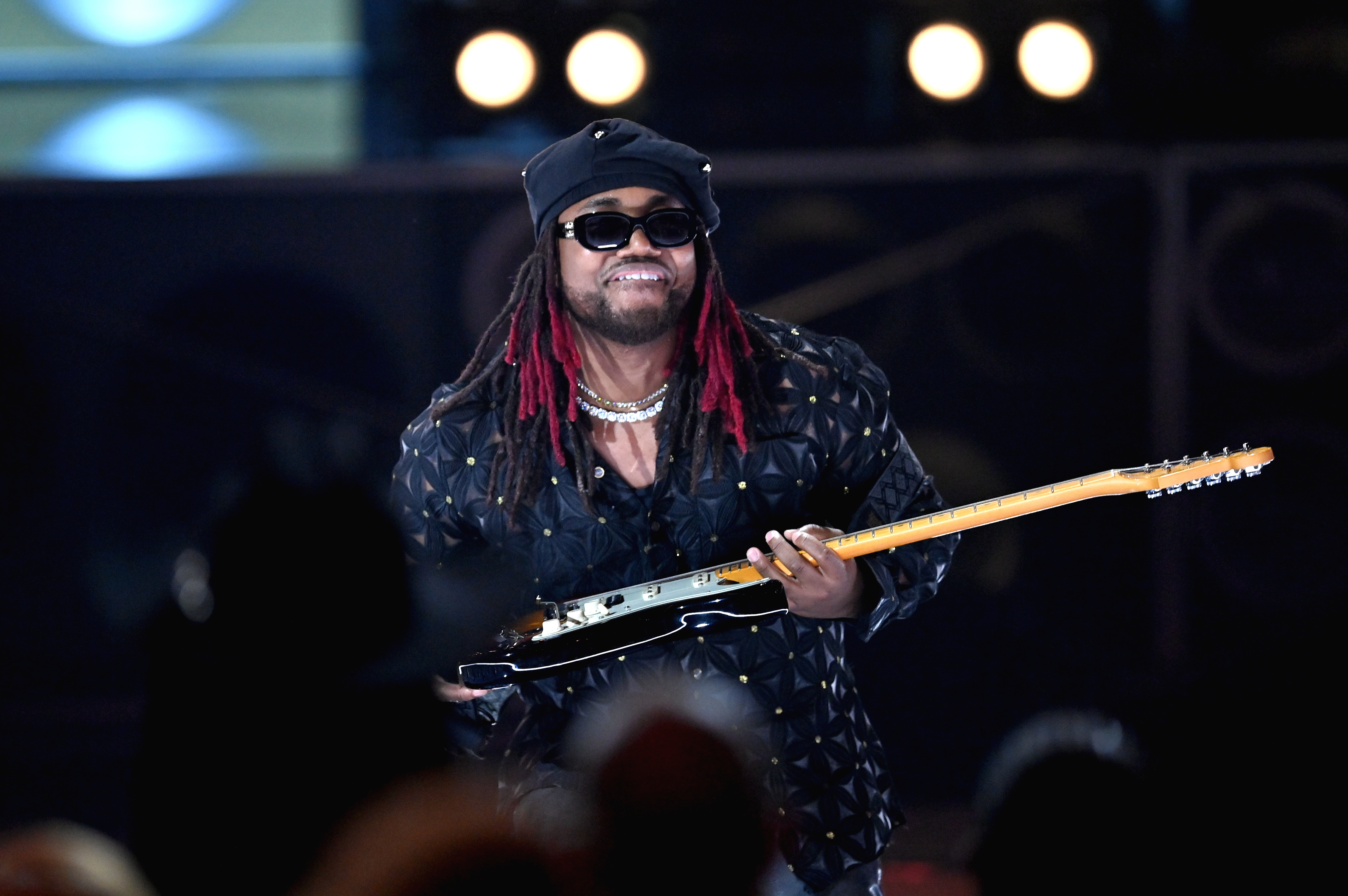 Leon Thomas, with red-tinted dreadlocks, sunglasses, and a hat, playing an electric guitar on stage with bright lights in the background. He was up for a number of Grammys. 