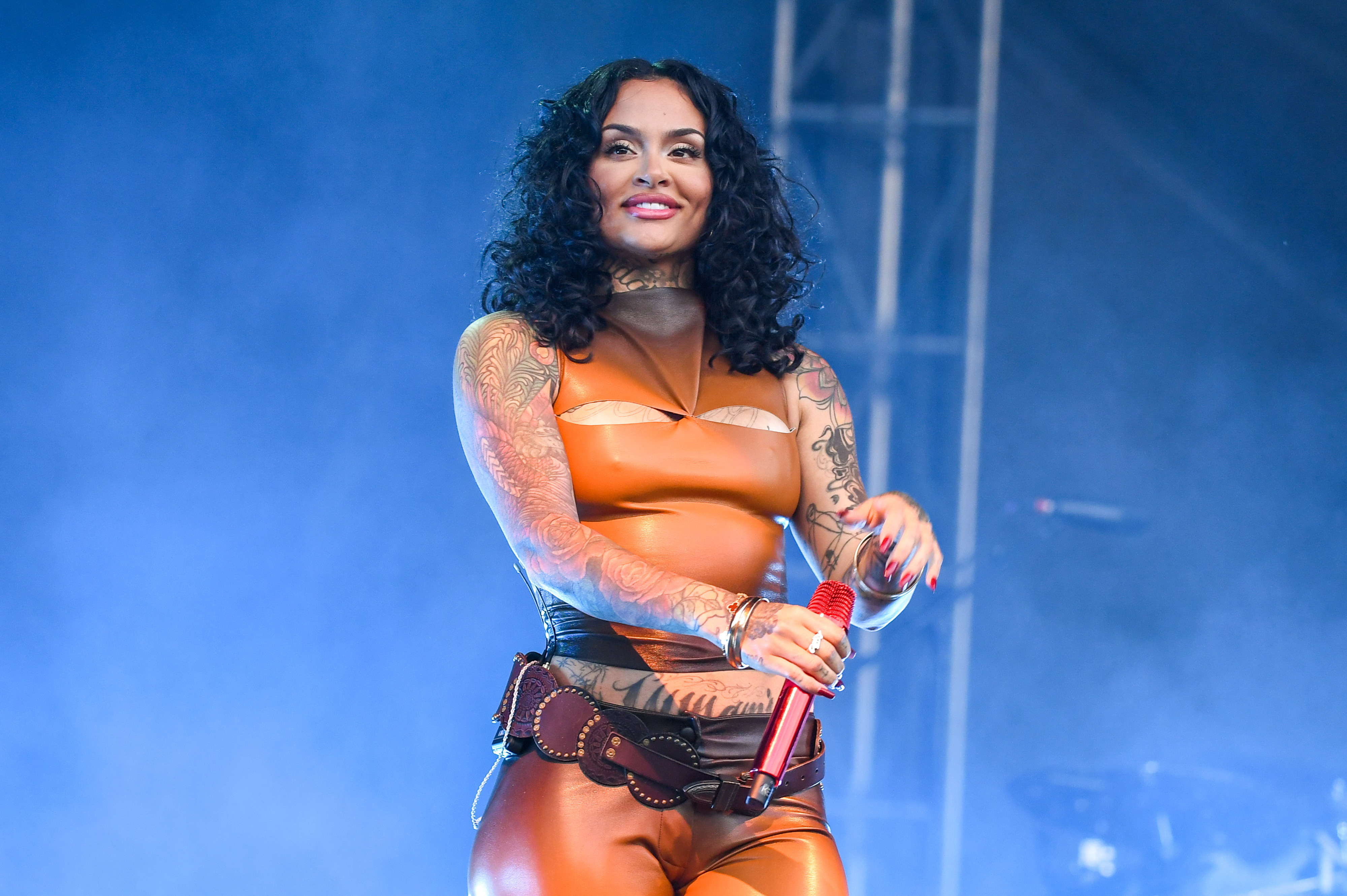 Kehlani with curly hair and tattoos performs on stage in a brown outfit, holding a red microphone. She might win her first Grammy for 