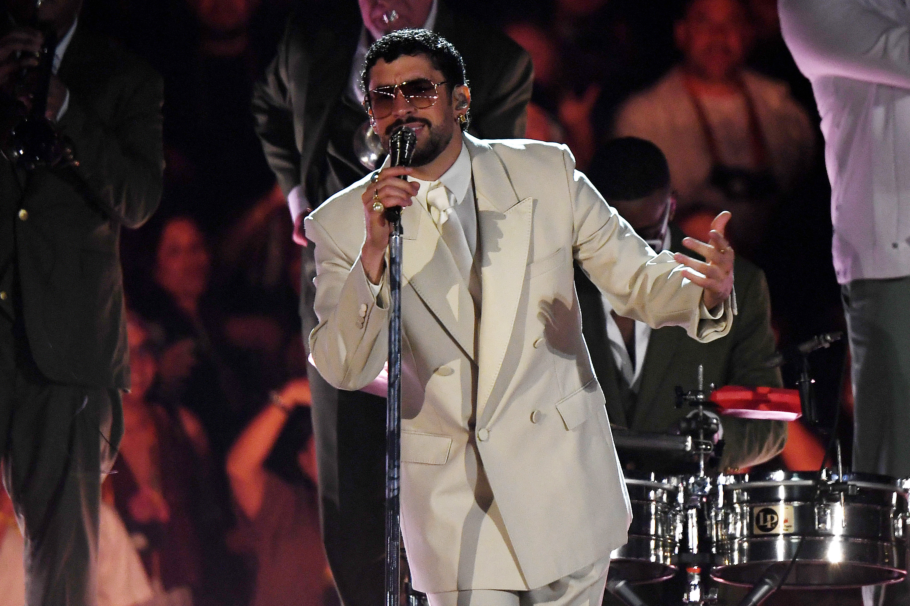 Bad Bunny in a white suit and sunglasses performs on stage, singing into a microphone, with musicians and a lively audience in the background. He is expected to win big at the Grammys