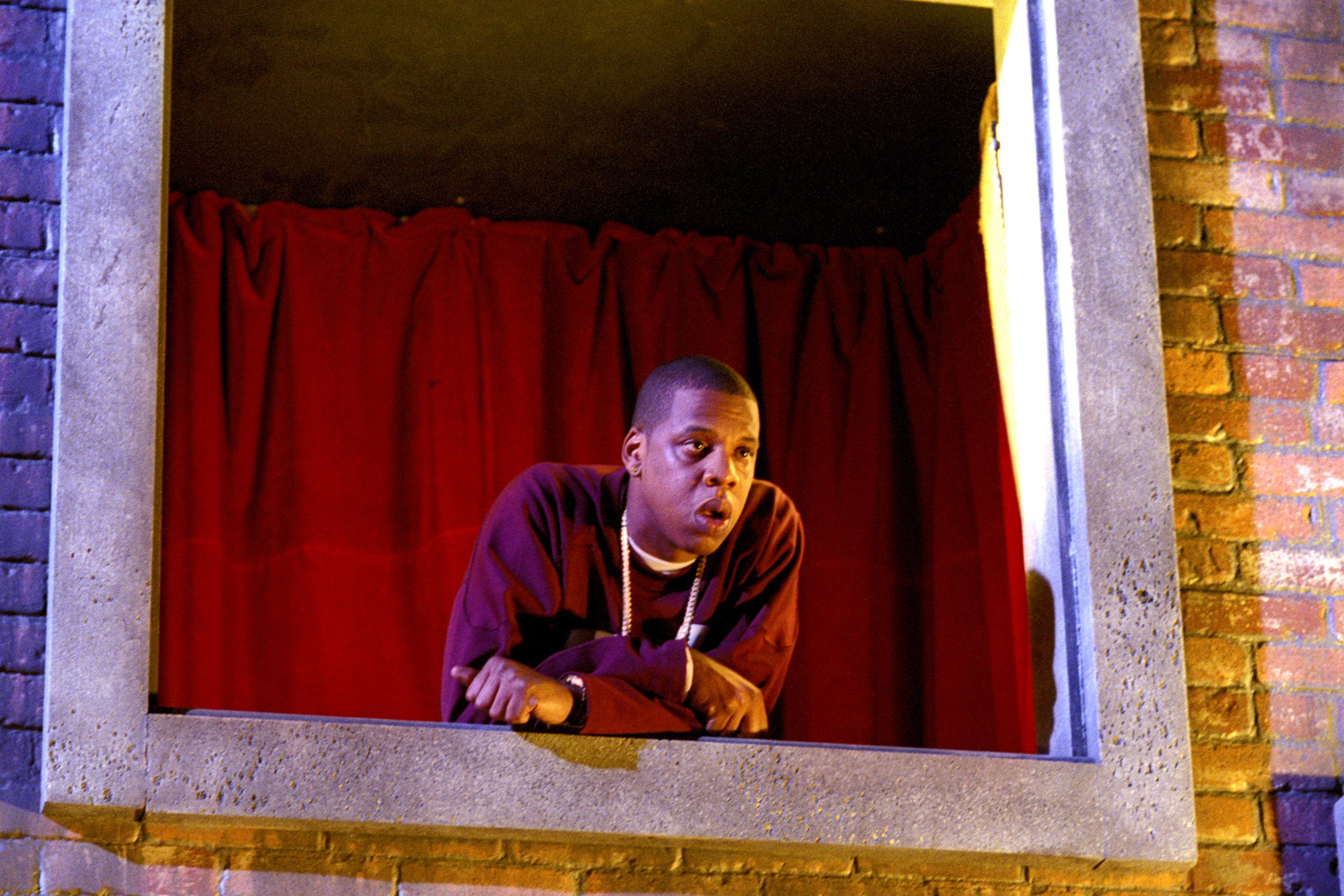Jay-Z leaning out of a window with red curtains, wearing a dark jacket and necklace, against a brick wall backdrop. This is from the 