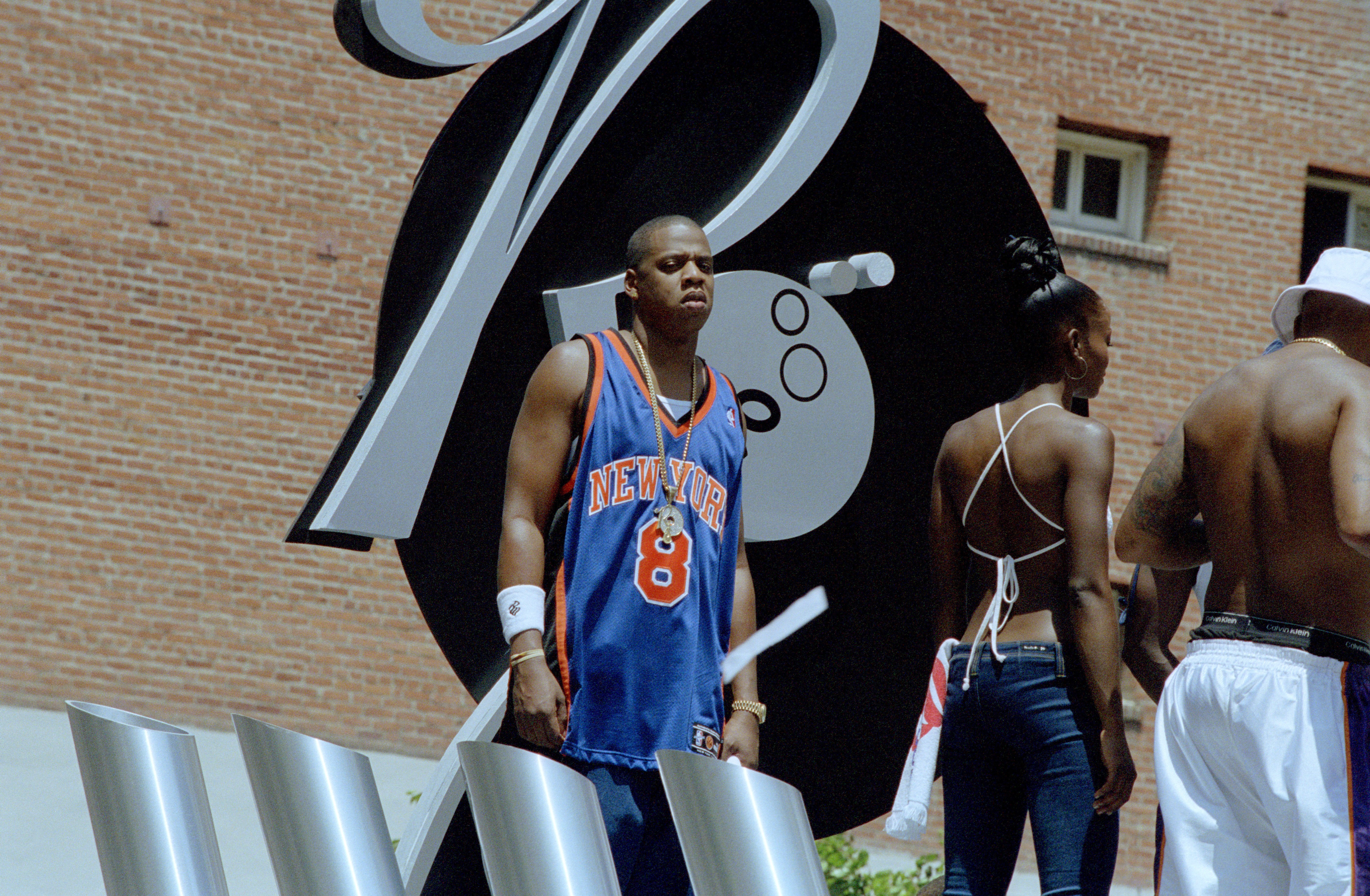 Jay-Z a New York Knicks jersey stands in front of a large black and white sculpture. Two other people are nearby. This is from the 
