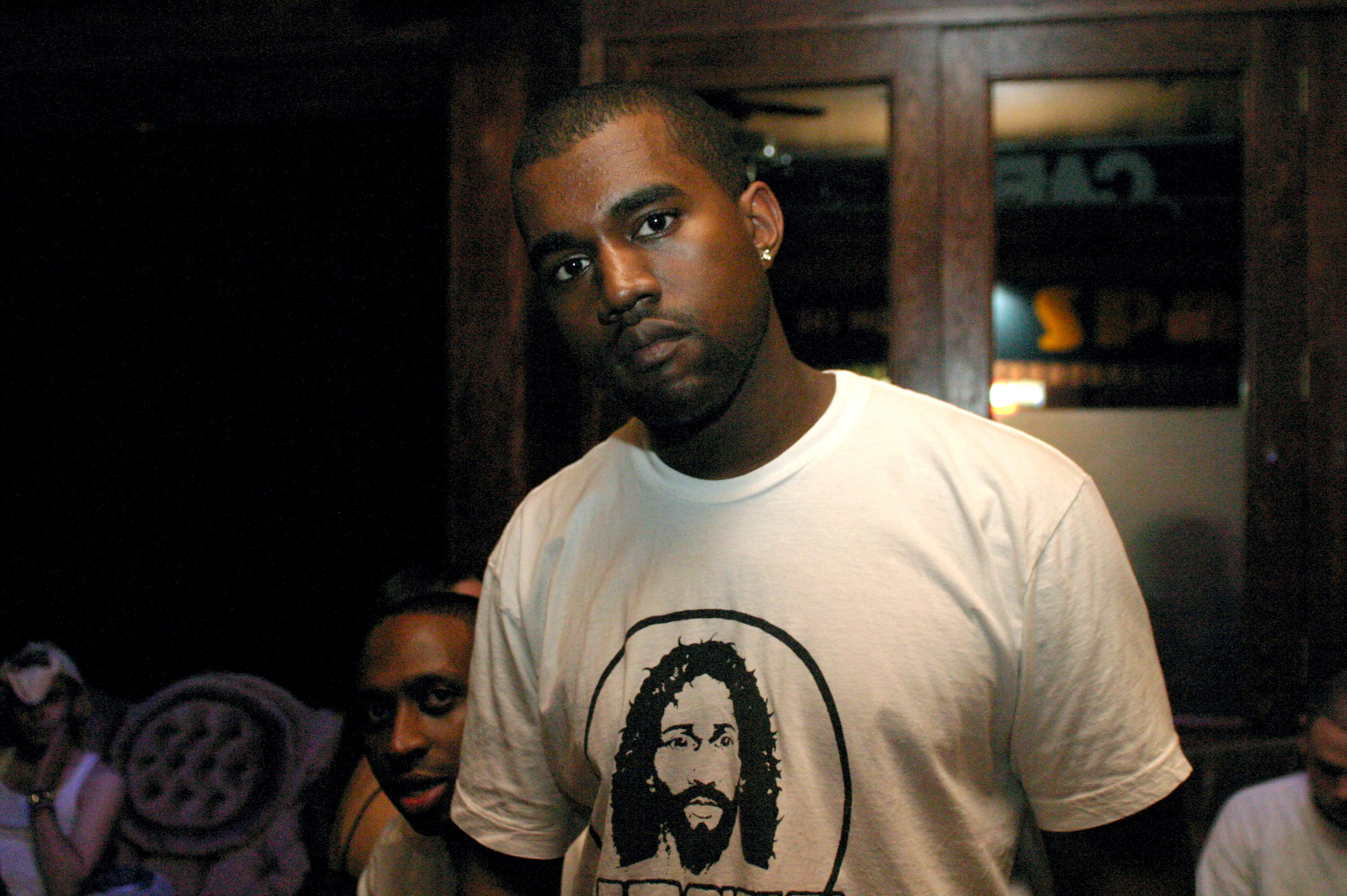 Kanye West wearing a white T-shirt with a graphic, standing indoors with people in the background. He was one of the main producers on The Blueprint 