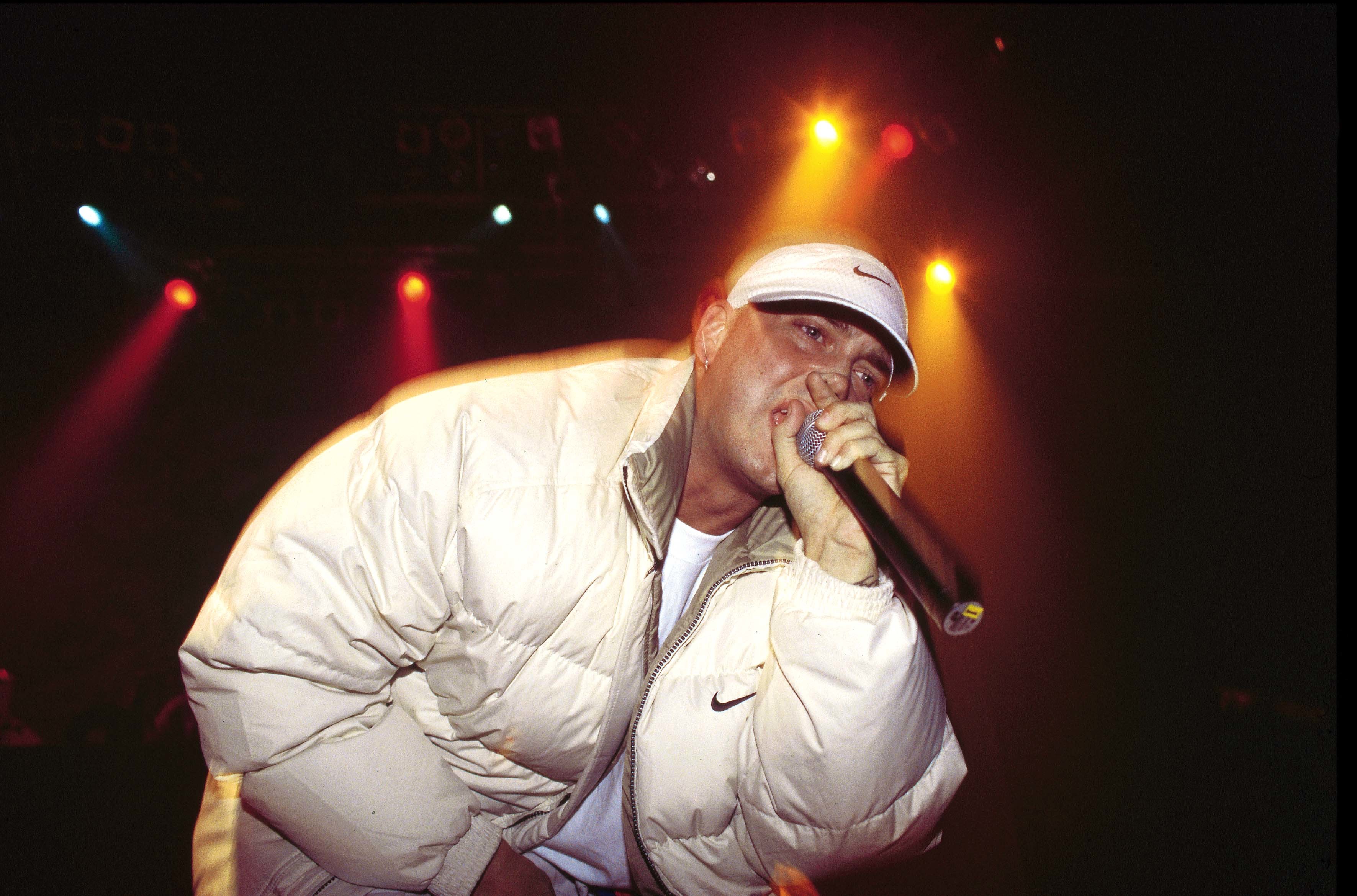 Rapper Eminem performing on stage, wearing a white jacket and cap, holding a microphone under colorful stage lights. He was one of the best verses on Jay-z's 'The Blueprint.'