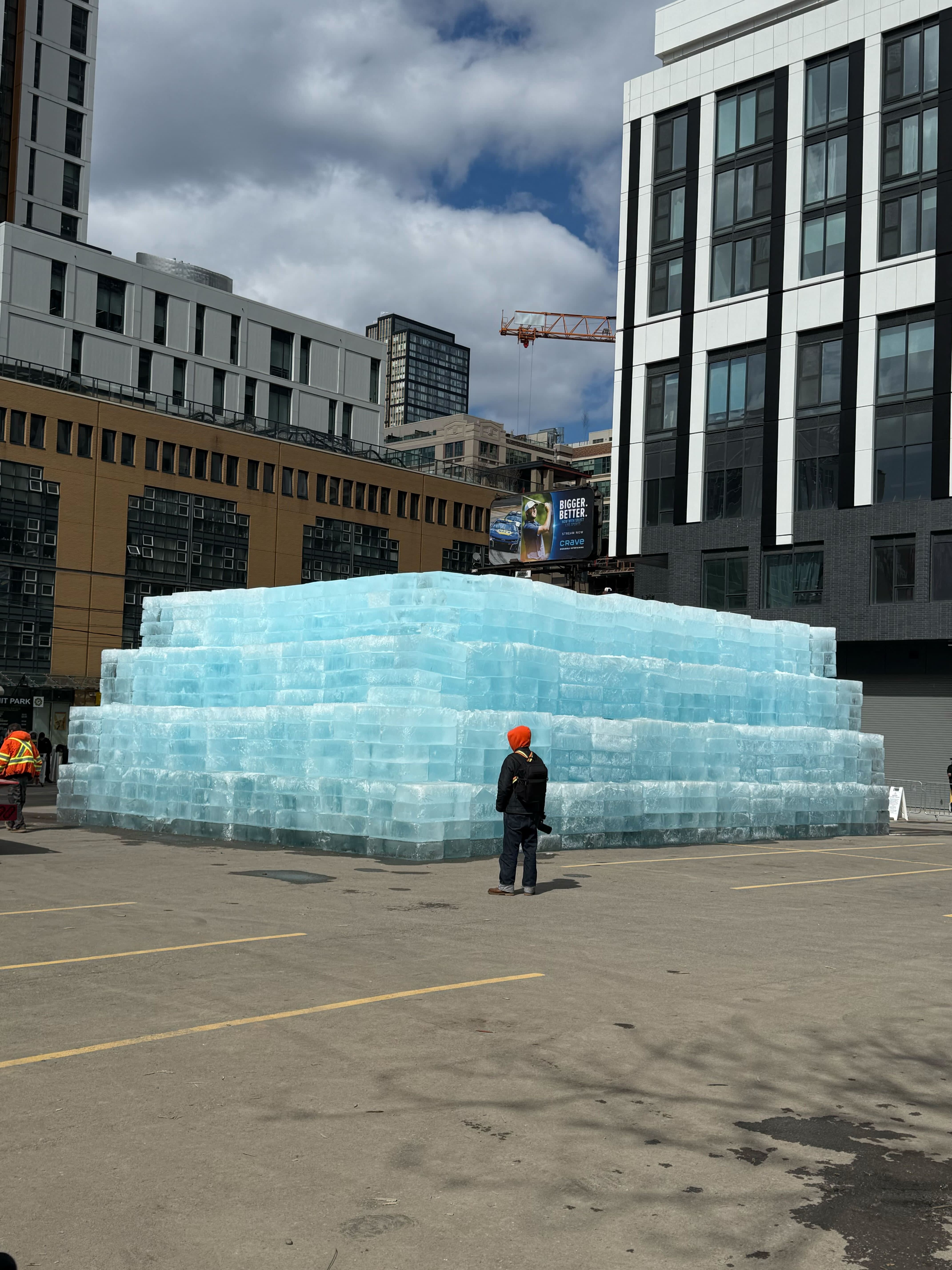 A large pyramid of ice blocks in an urban setting, with two people in orange hats nearby. Buildings and a crane in the background.
