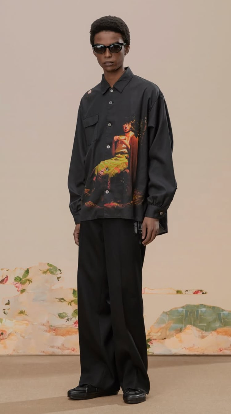 A man wears an all-black outfit with an orange graphic on a button-up long sleeve shirt.