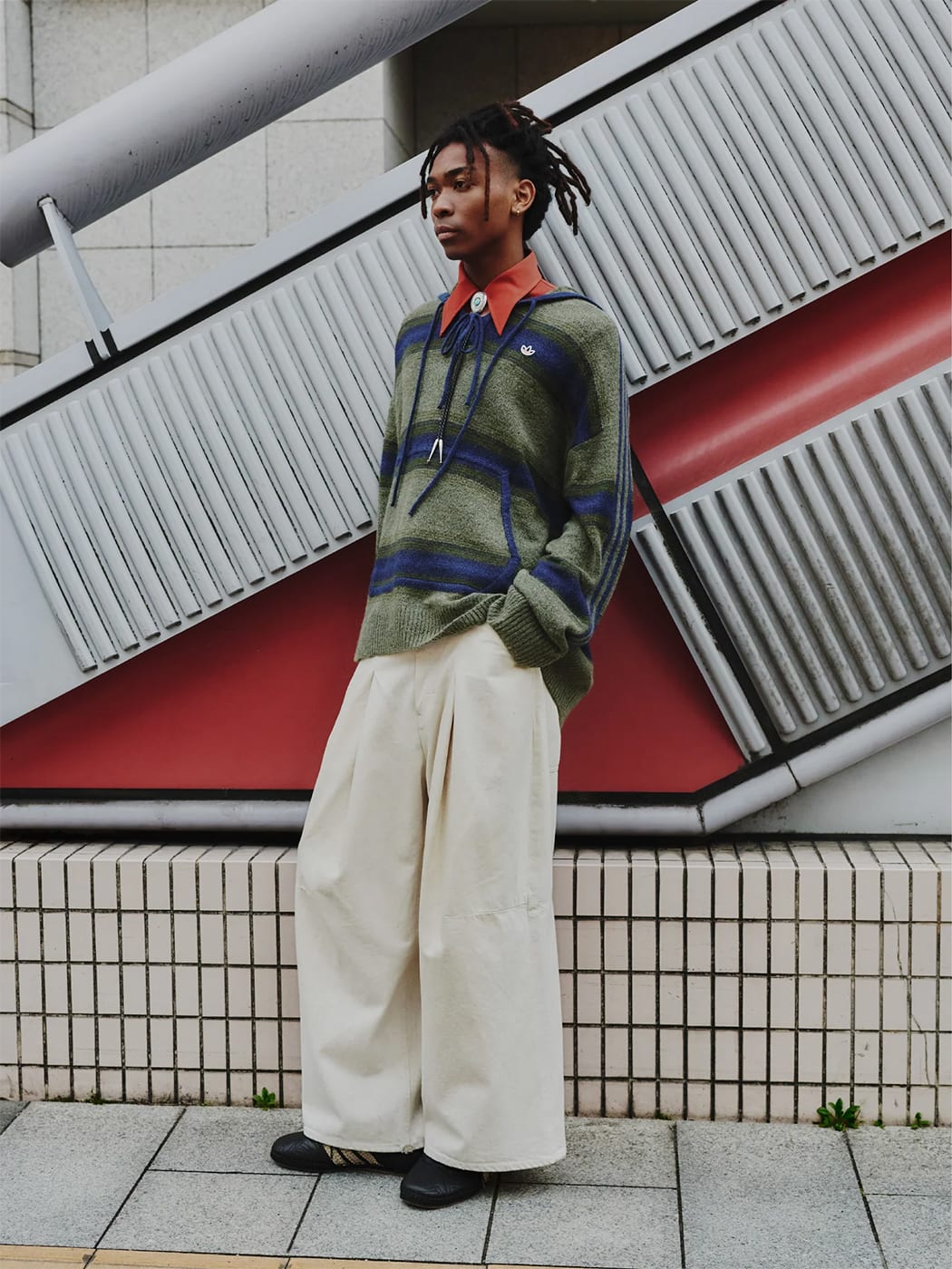 A man wears a collared red shirt under a blue and yellow striped sweater paired with white, wide-leg pants and black shoes.